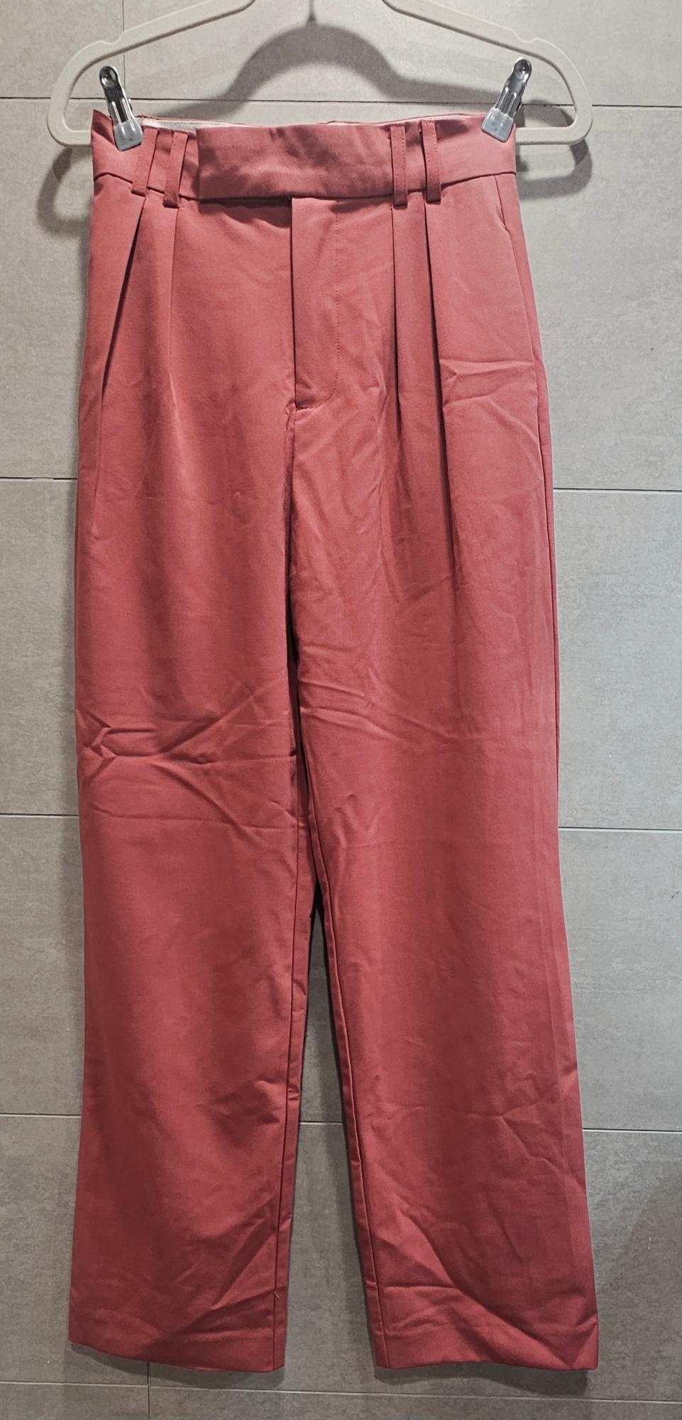 Dark pink trousers hanging on a hanger against a grey tiled background.