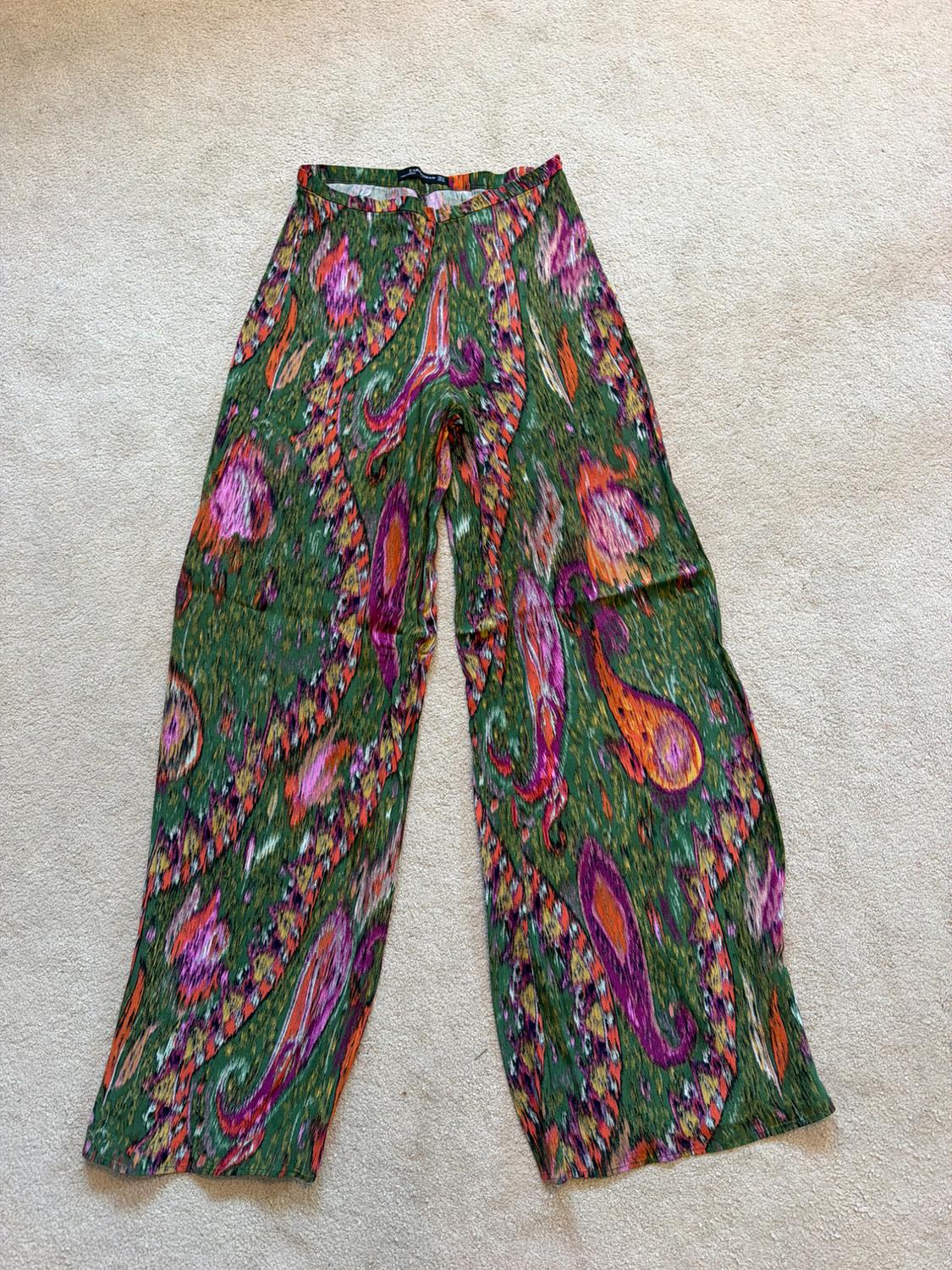 Wide-leg trousers with an abstract print in green, pink, and orange tones, on a beige rug.