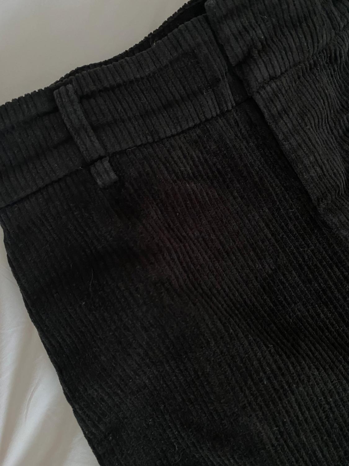 Close-up of black corduroy trousers showing waistband detail with belt loops on a white fabric.