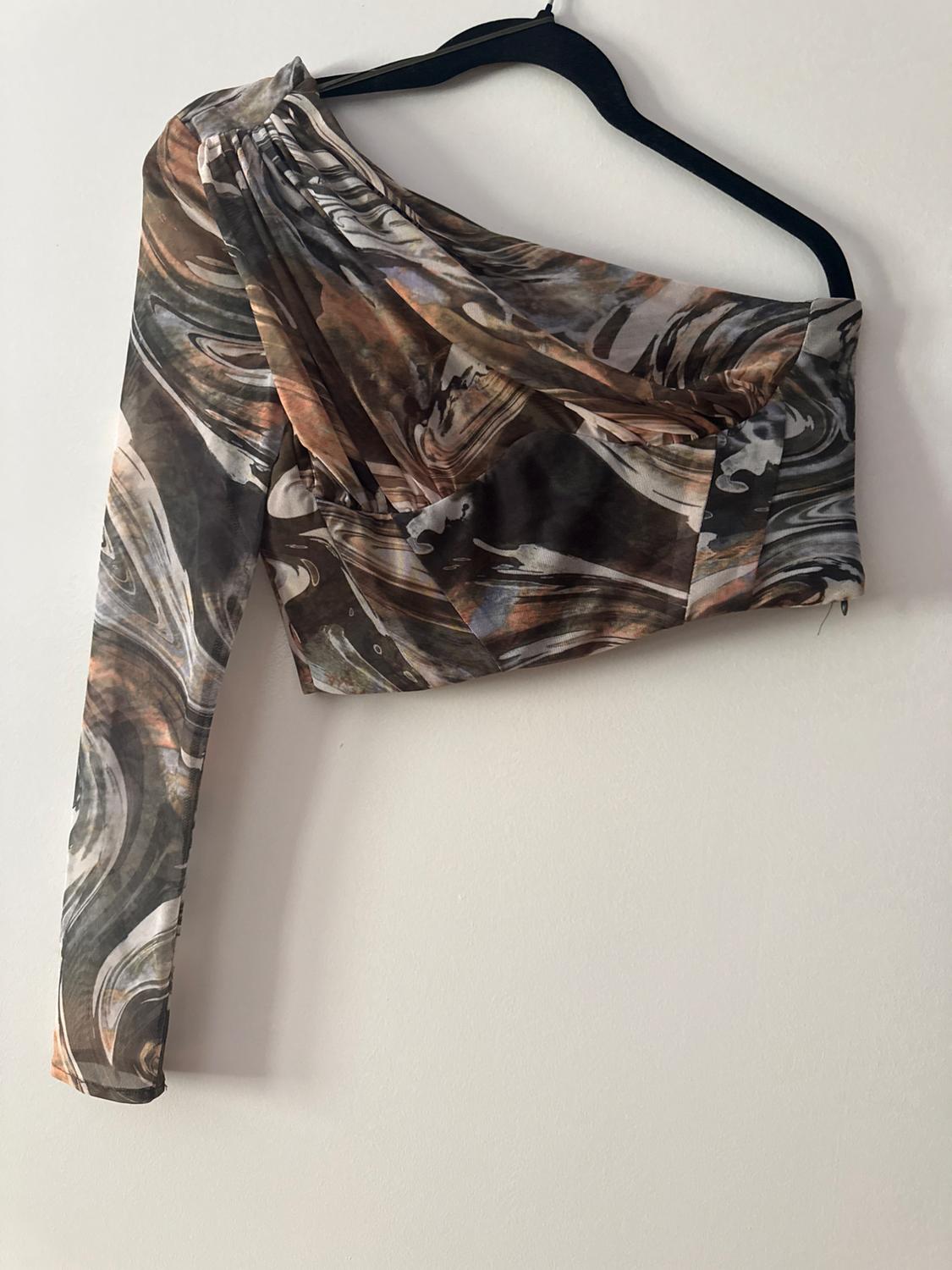 Asymmetric one-shoulder crop top with an abstract print in earthy tones and black, hanging on a black hanger against a white wall.