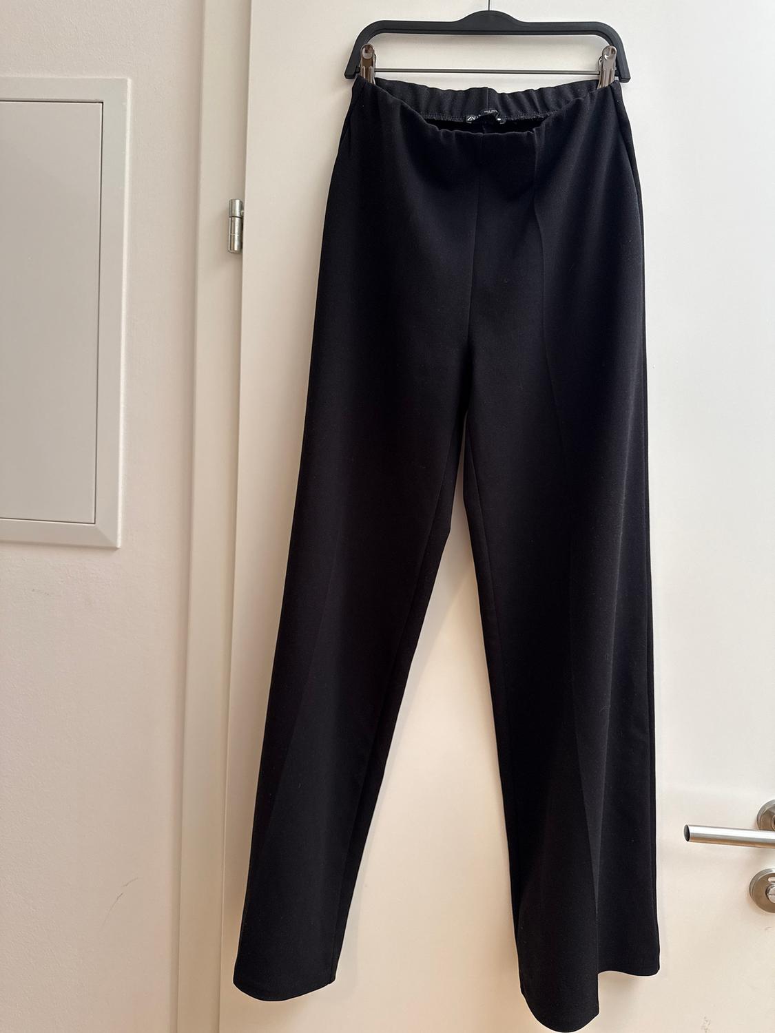 Black trousers hanging on a hanger over a white door. The fabric looks soft and the design is simple and elegant.