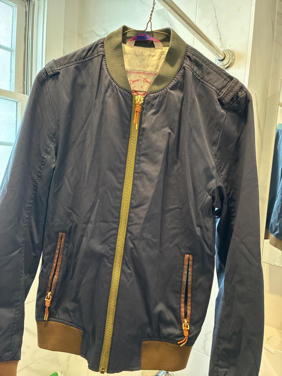Navy blue bomber jacket on a hanger. It has a gold zipper, green collar, brown hem, and pockets. Inner label: “Denim Couture.”