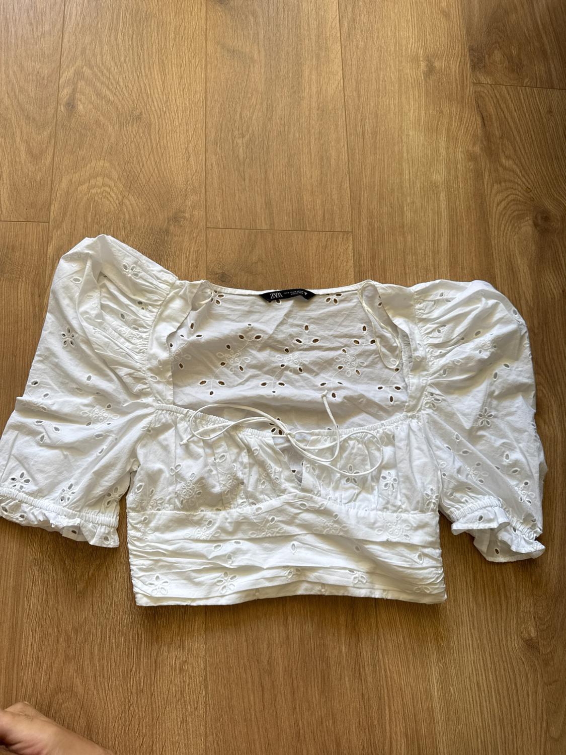 White cropped top with broderie anglaise, puff sleeves and a front tie, laid out on a wooden floor. ZARA label.