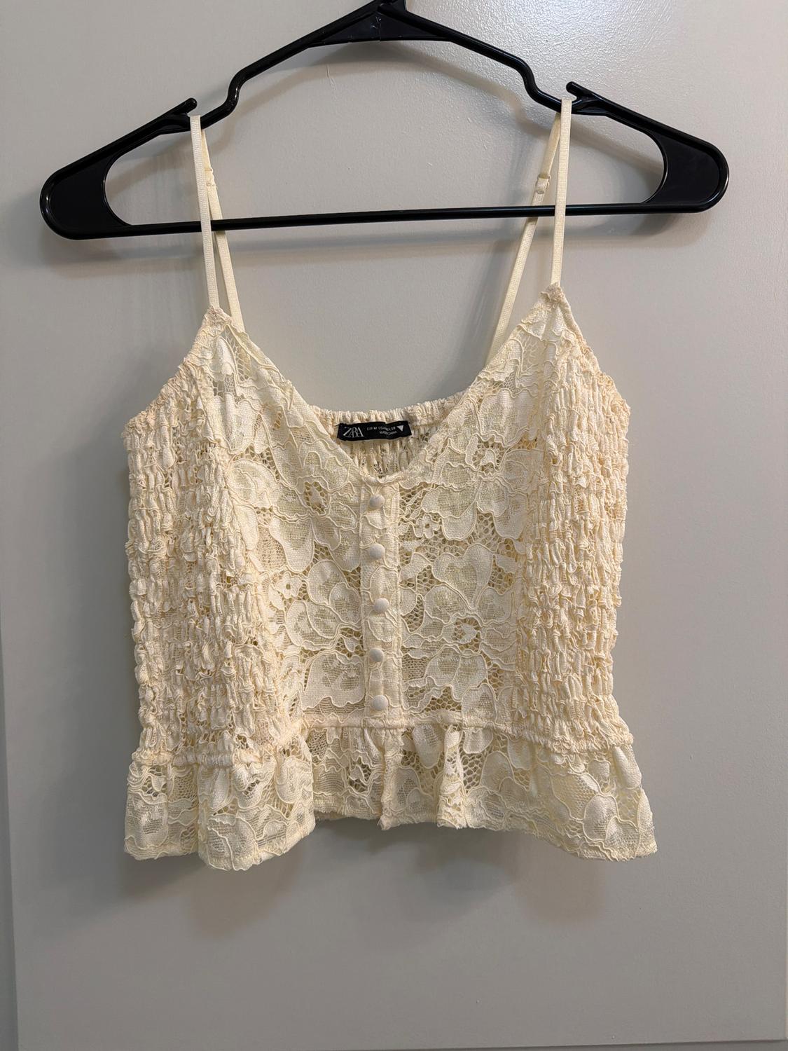 Beige lace top with thin straps and front buttons, hanging on a black hanger against a gray wall. Visible tag: Zara.