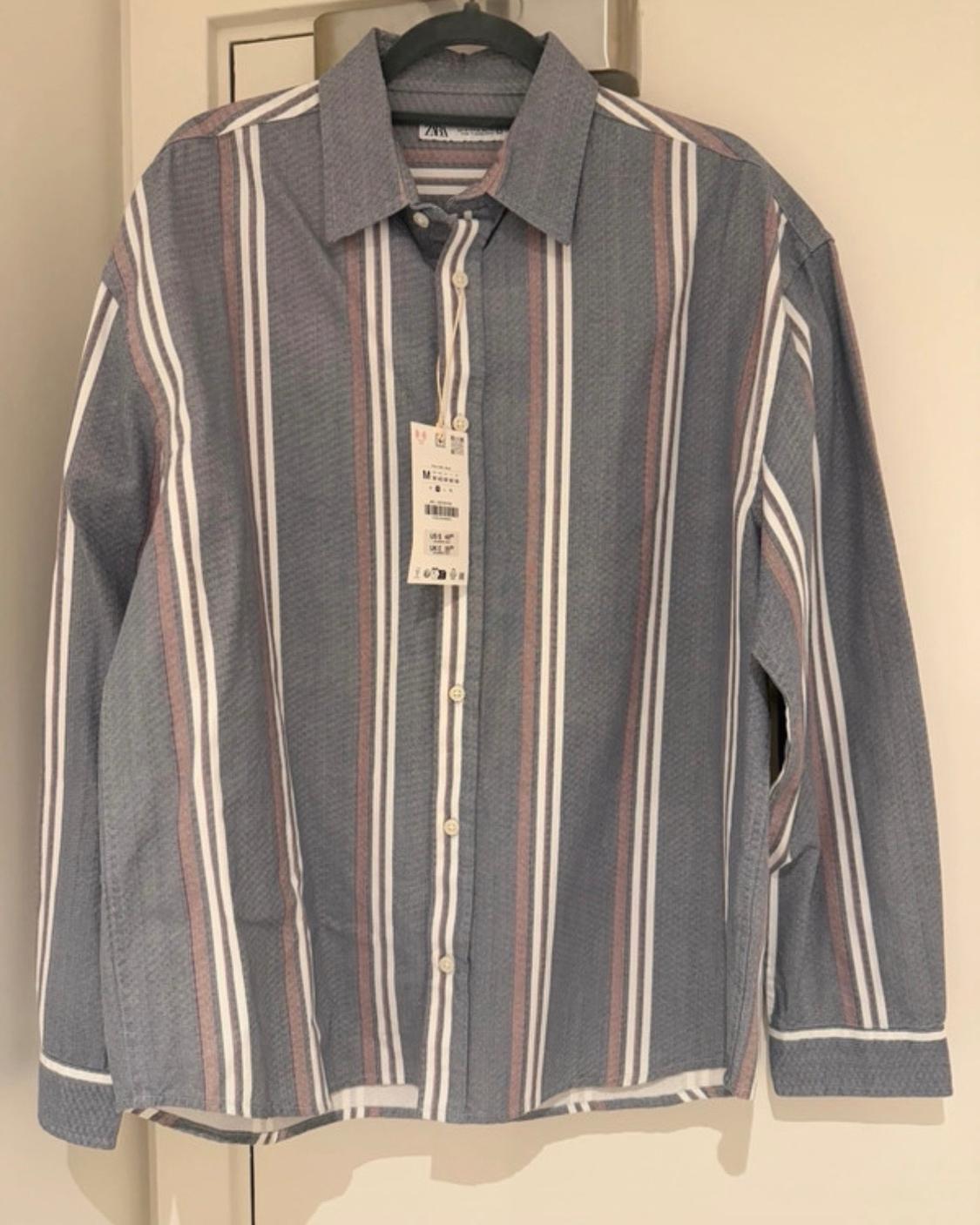 Greyish-blue ZARA long-sleeved shirt with white and red stripes. It has a size M label and is hanging from a hanger on a white door.