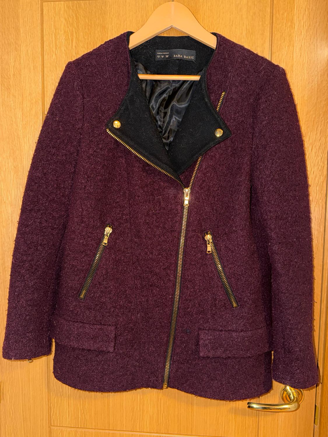 Burgundy jacket with a gold zip and pockets, hanging on a wooden hanger over a light wooden door. Label: ZARA BASIC.