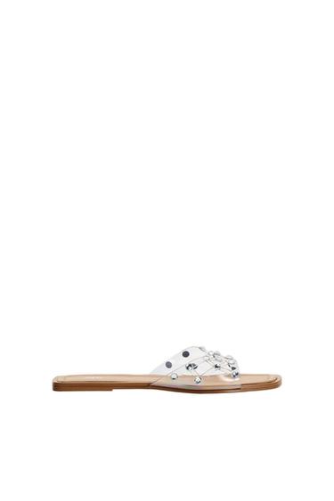 Flat sandal with a brown sole and a transparent strap decorated with silver studs and black dots on a white background.