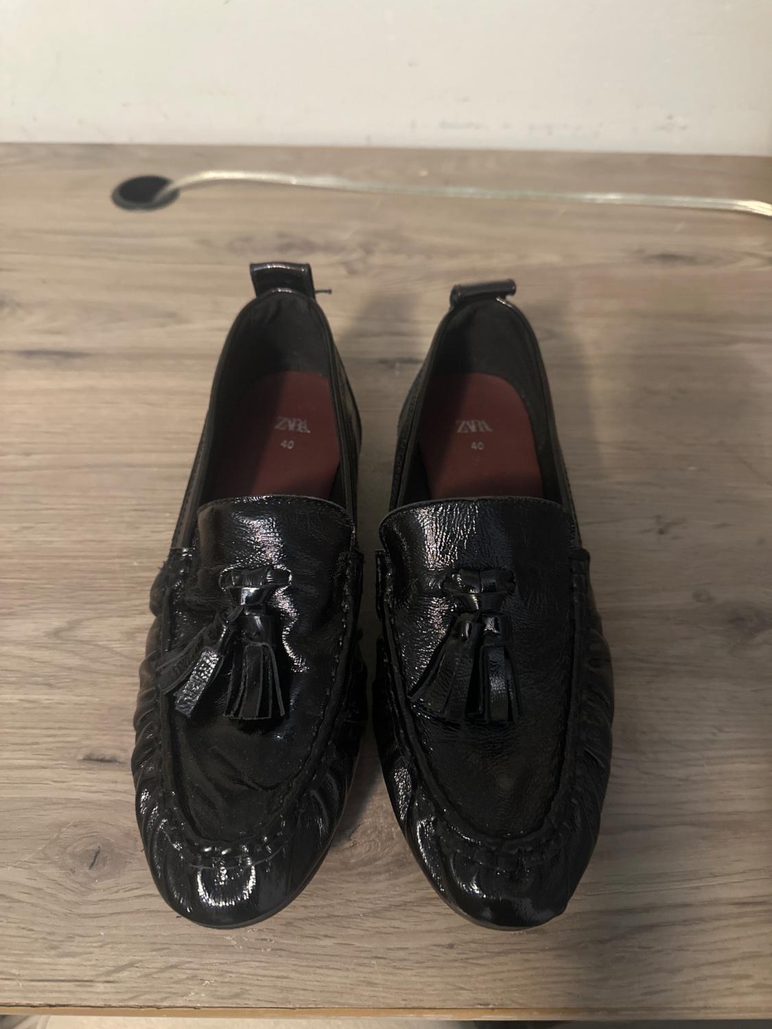 Black patent leather loafers with tassels on a light wooden surface. Visible text: "ZARA 40".