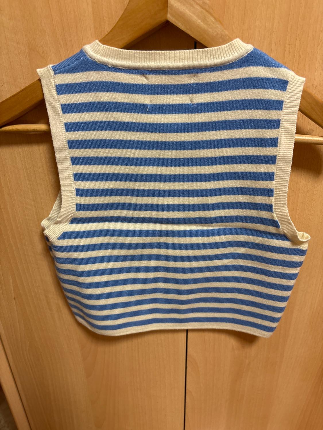 Knitted waistcoat with blue and white horizontal stripes, hanging on a wooden hanger in front of a wooden wardrobe.