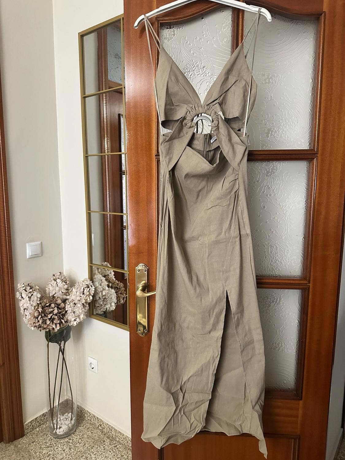 Beige dress with cut-outs and a central ring hanging on a wooden door. Next to it, a wall mirror and a vase with dried flowers.