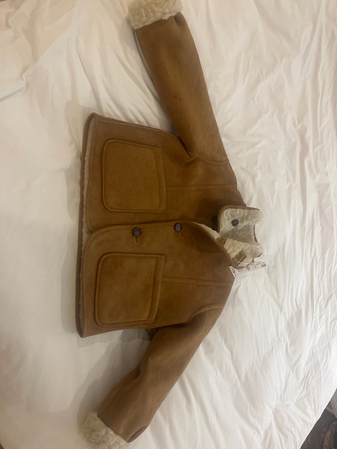 A brown faux leather jacket with a wool lining, large pockets, and dark buttons, on a white bed.