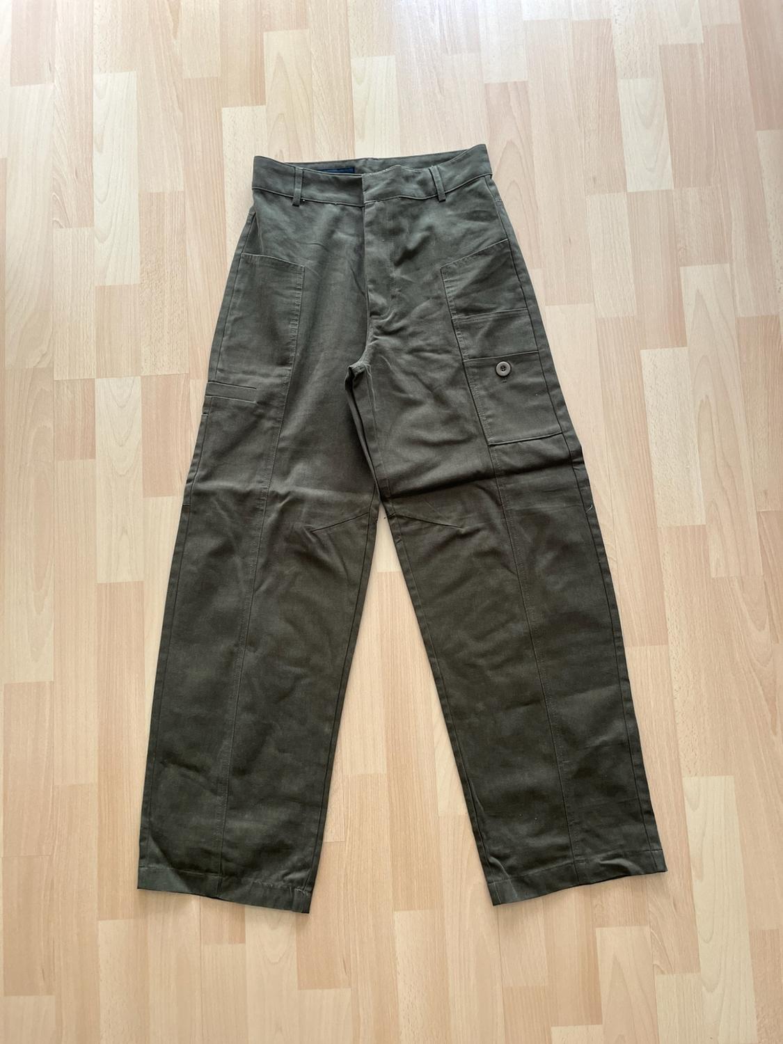 Wide olive green trousers with large front pockets, one with a button, and prominent stitching, laid on a light wooden floor.