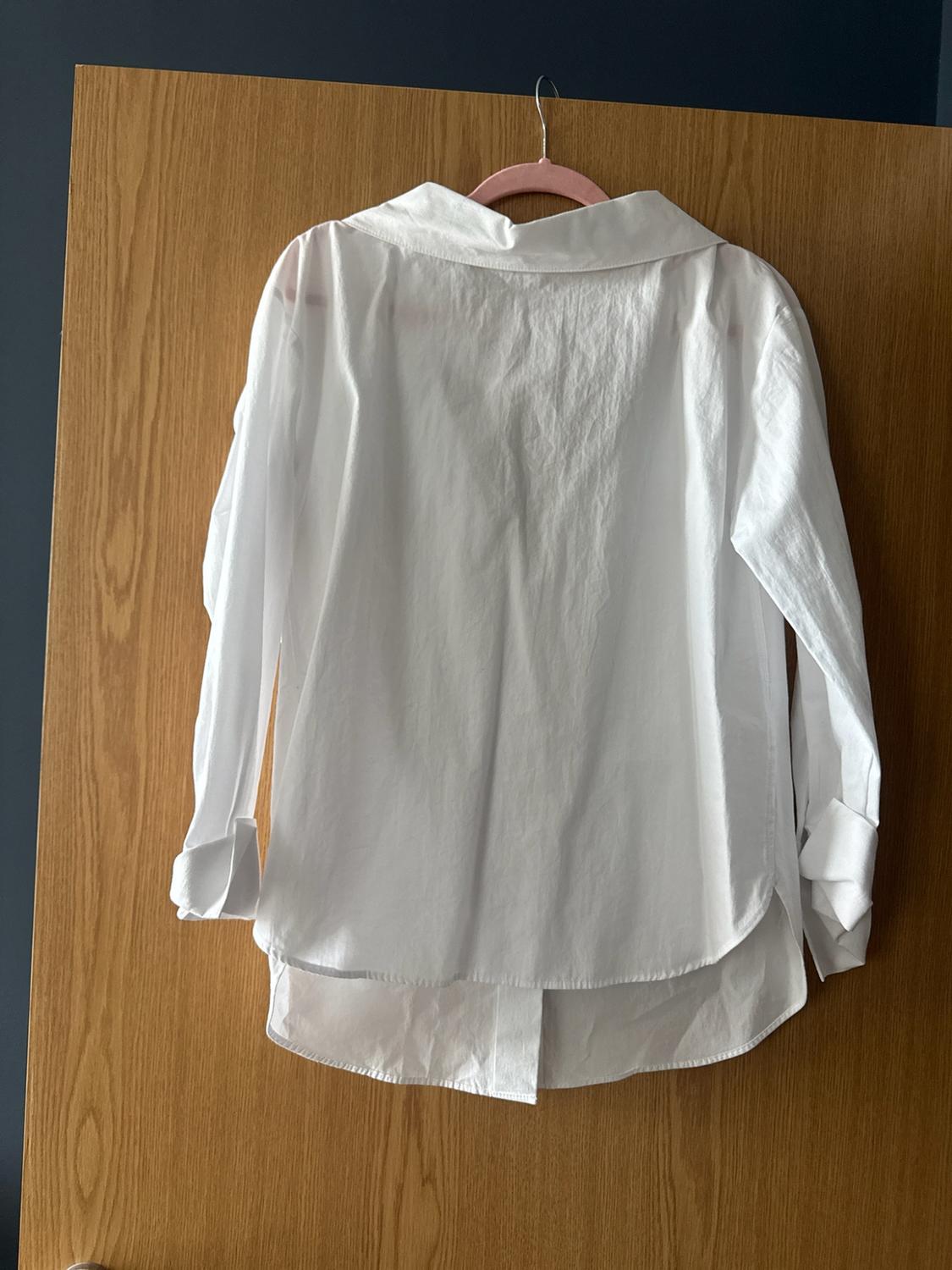 Long-sleeved white shirt seen from behind, hanging on a pink hanger on a wooden door. It features a collar and an asymmetrical hem.
