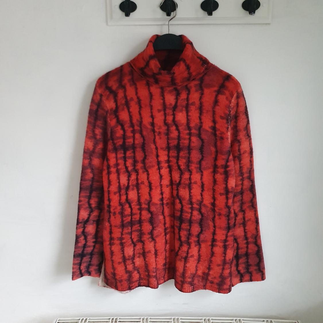 Red jumper with black stripe pattern hanging on a hanger against a white wall.