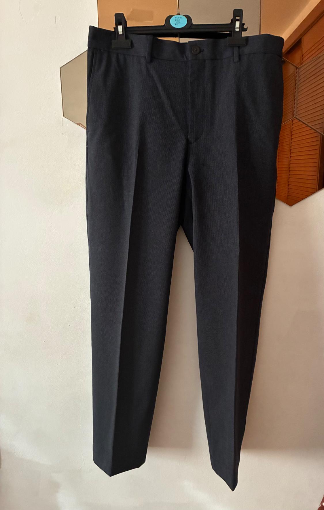 Black trousers hanging on a black hanger with clips, against a light background and a wooden-panelled wall.