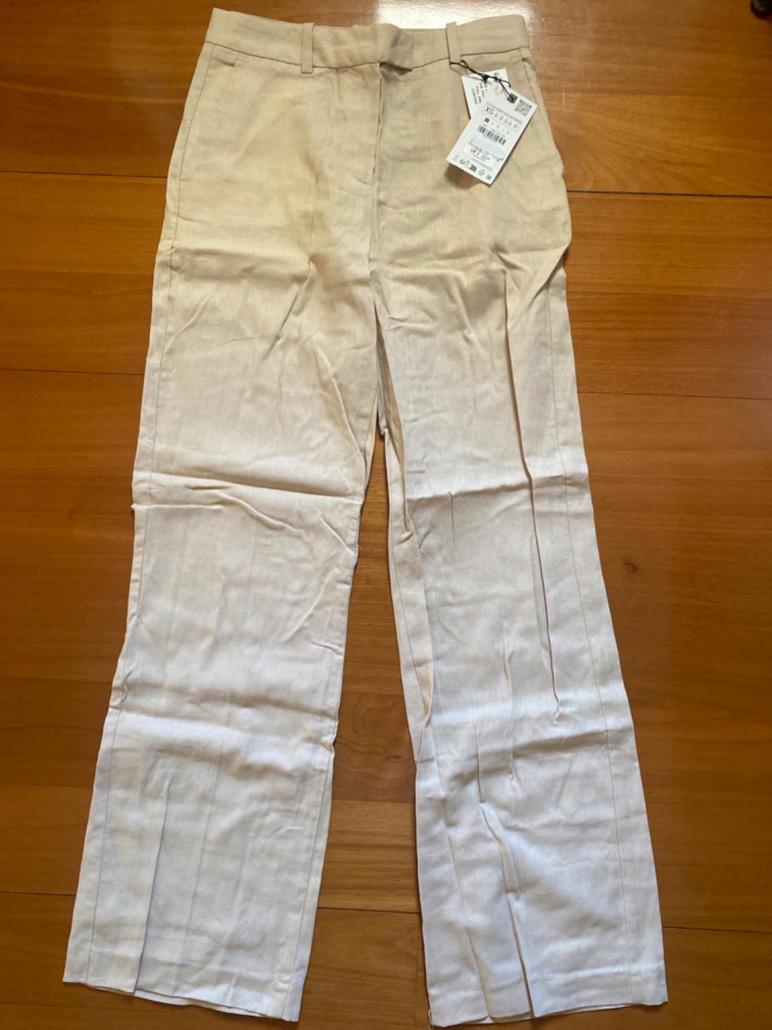 Beige linen trousers with a swing tag, on a wooden floor.
