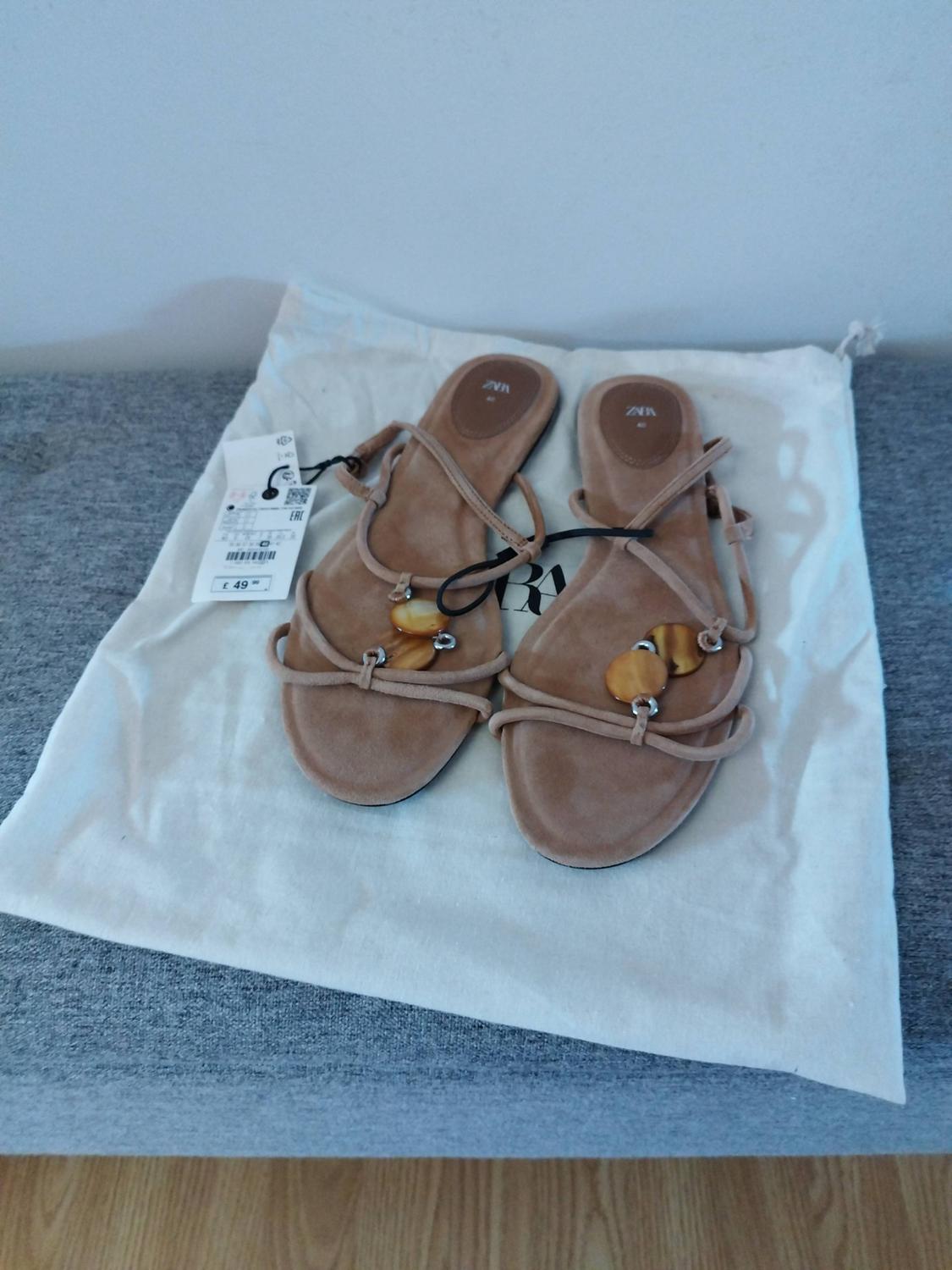 Beige strappy flat sandals with embellishments on a white fabric bag on a grey surface. Text: ZARA, EAC, £ 49.99.