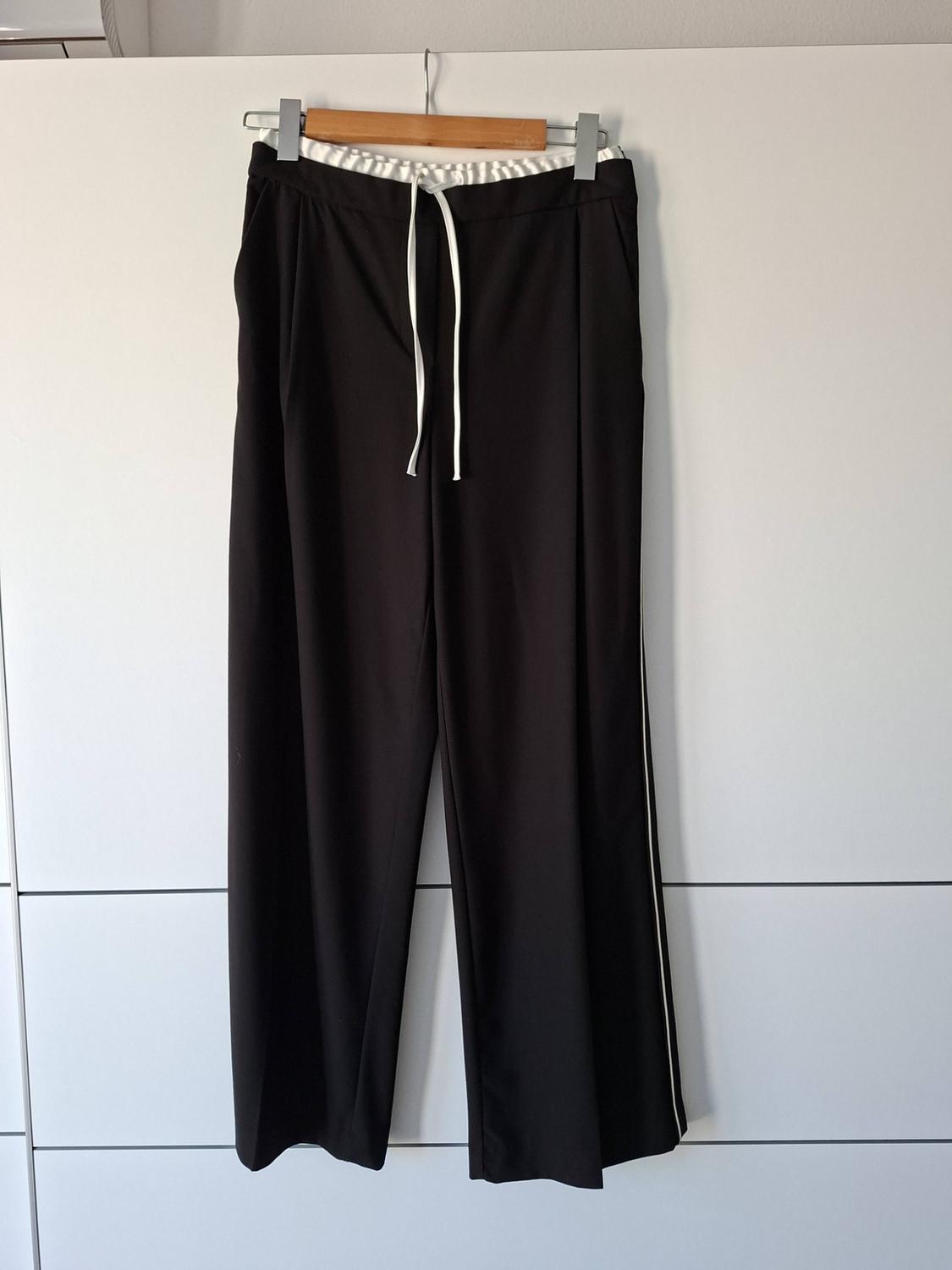 Wide-leg black trousers with a white drawstring and a fine side stripe, hanging on a wooden hanger above a white wardrobe.