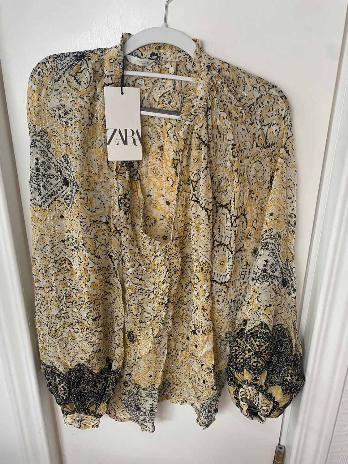 Printed blouse in yellow and black tones hanging on a white hanger. Label with the text "ZARA" visible.