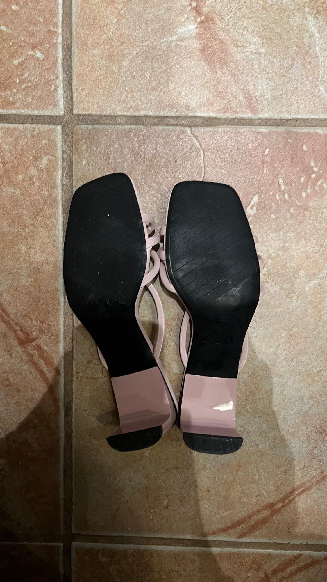 Pink block-heeled sandals with black soles, viewed from below on a brown tiled floor.