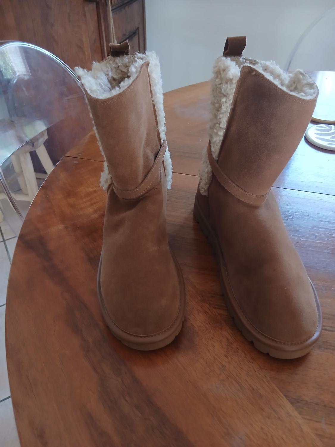 Brown suede shoes with a wool interior, placed on a wooden table. In the background, a transparent chair and a wooden cabinet are visible.