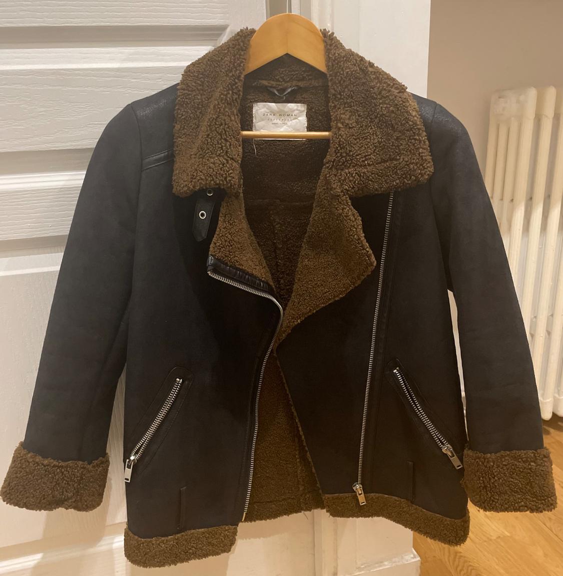Black jacket with brown sheepskin lining, zip fastening and pockets, hanging on a wooden hanger in front of a white door.