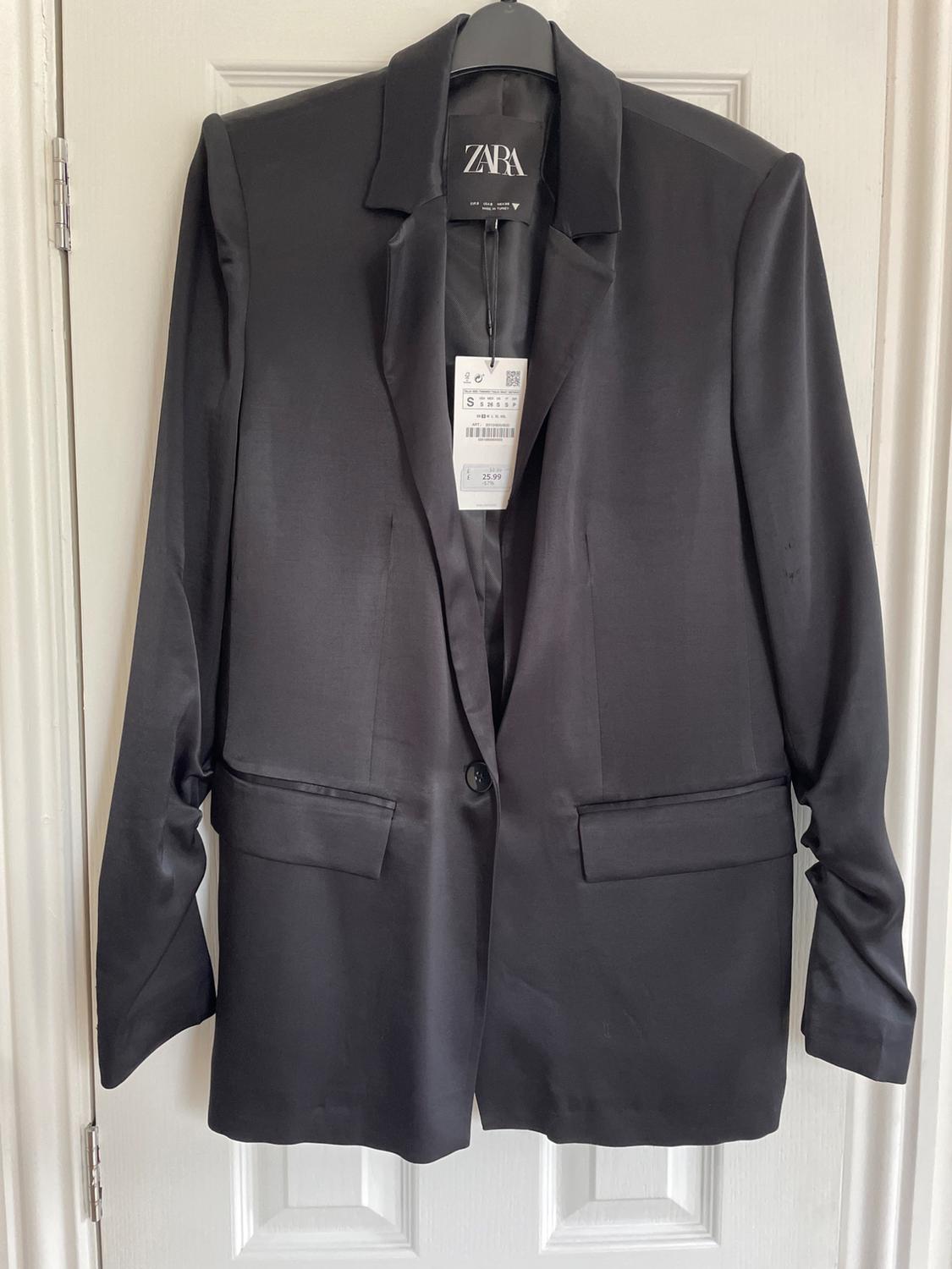 Black satin jacket hanging on a white door, with a Zara label visible on the inside.