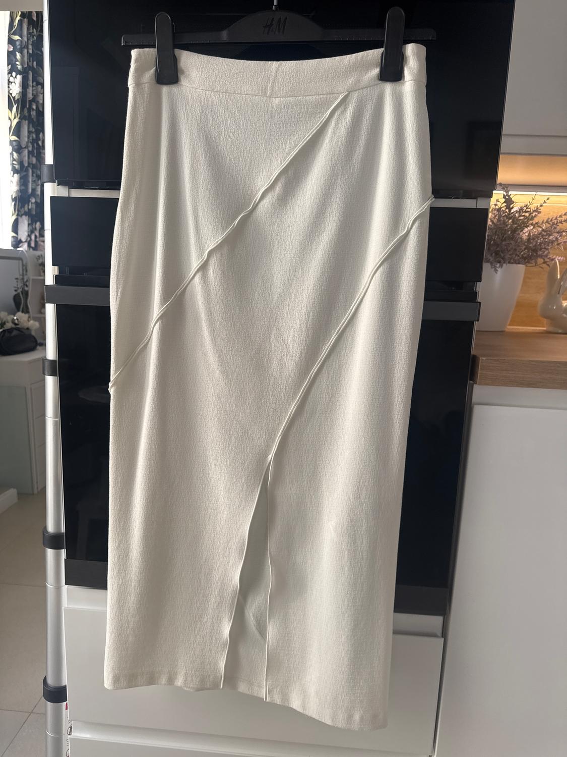 White skirt with a diagonal line design, hanging on a black H&M hanger. Modern kitchen background with floral decor.