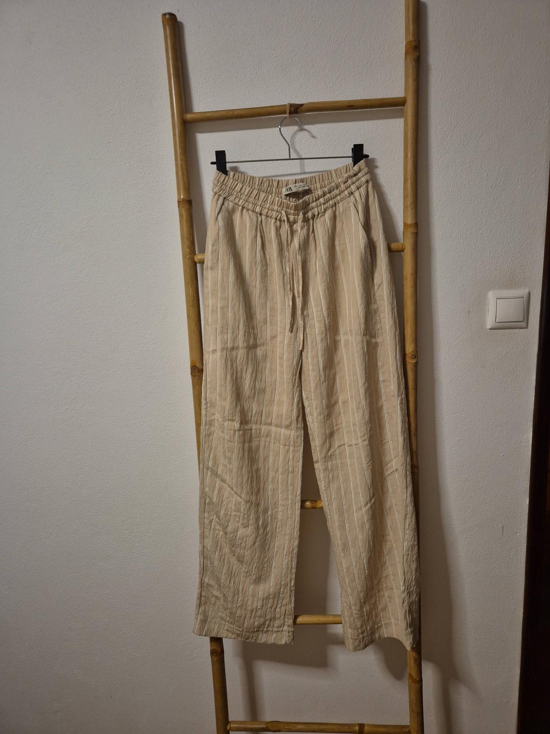 Beige trousers with white stripes and an elasticated waist with a drawstring, hanging on a wooden ladder leaning against a white wall.