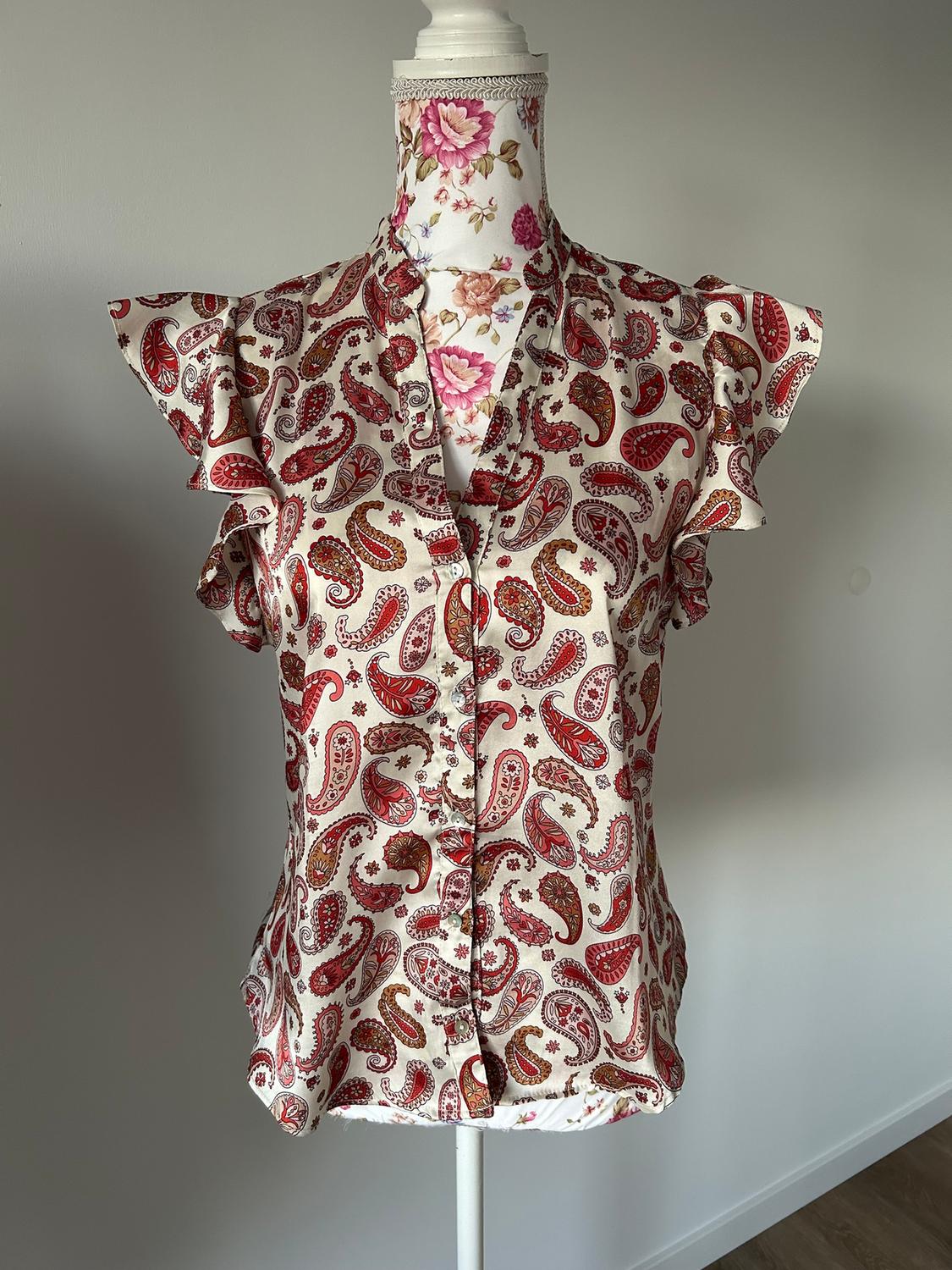 Short-sleeved blouse with a red and beige paisley print on a mannequin with a floral background.