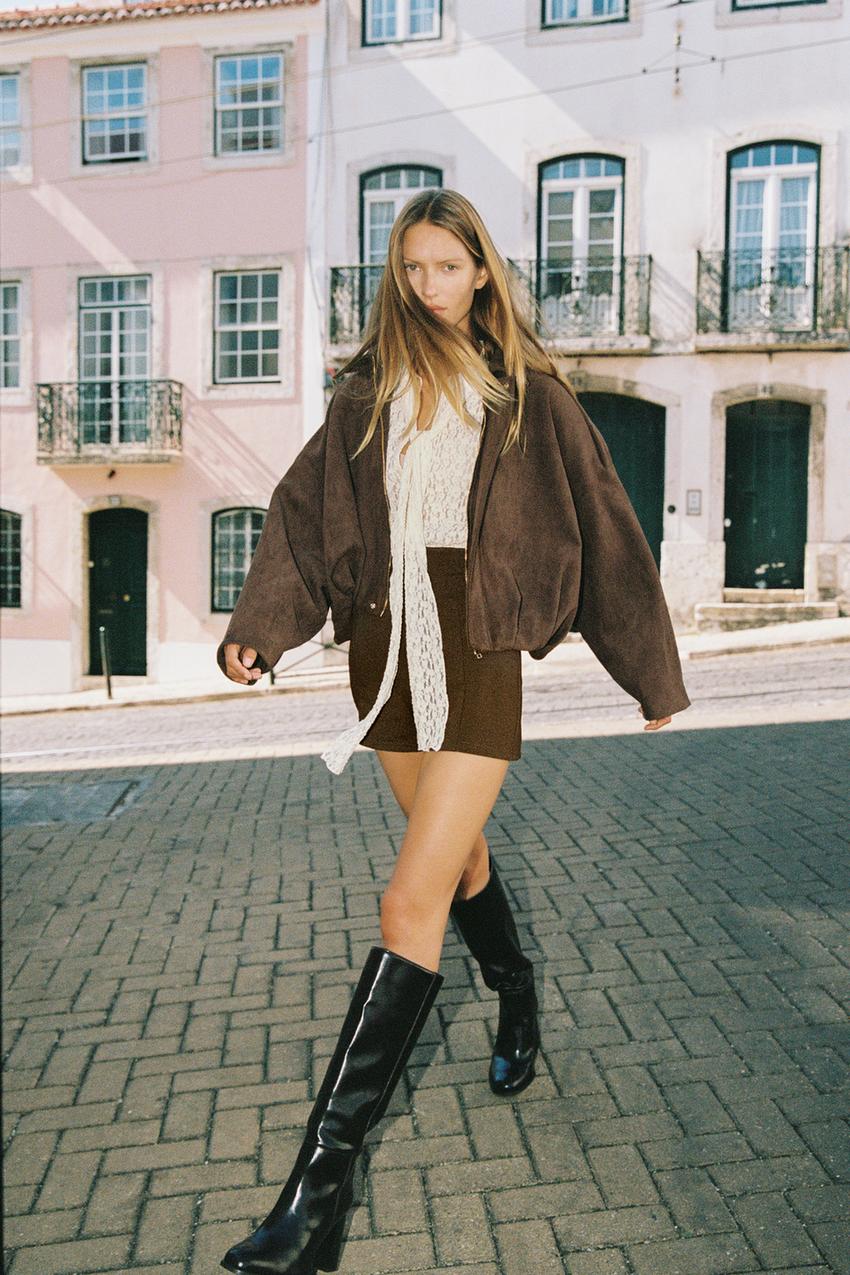 Dark brown bomber jacket set with a lapel collar and long sleeves, brown skirt, and black tall boots.
