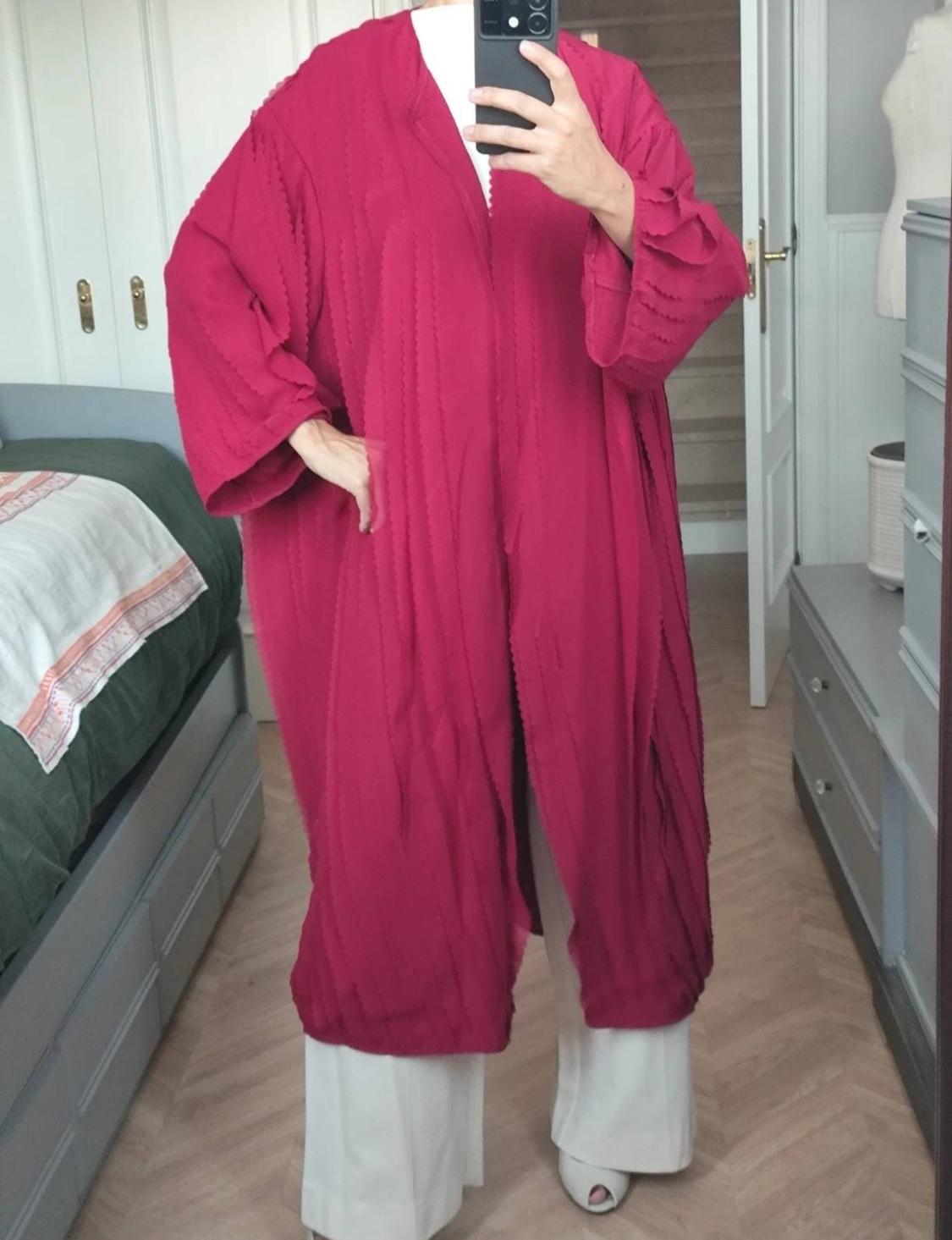 Person taking a selfie in the mirror of a room. They are wearing a textured fuchsia long coat, a white top and wide-leg trousers.