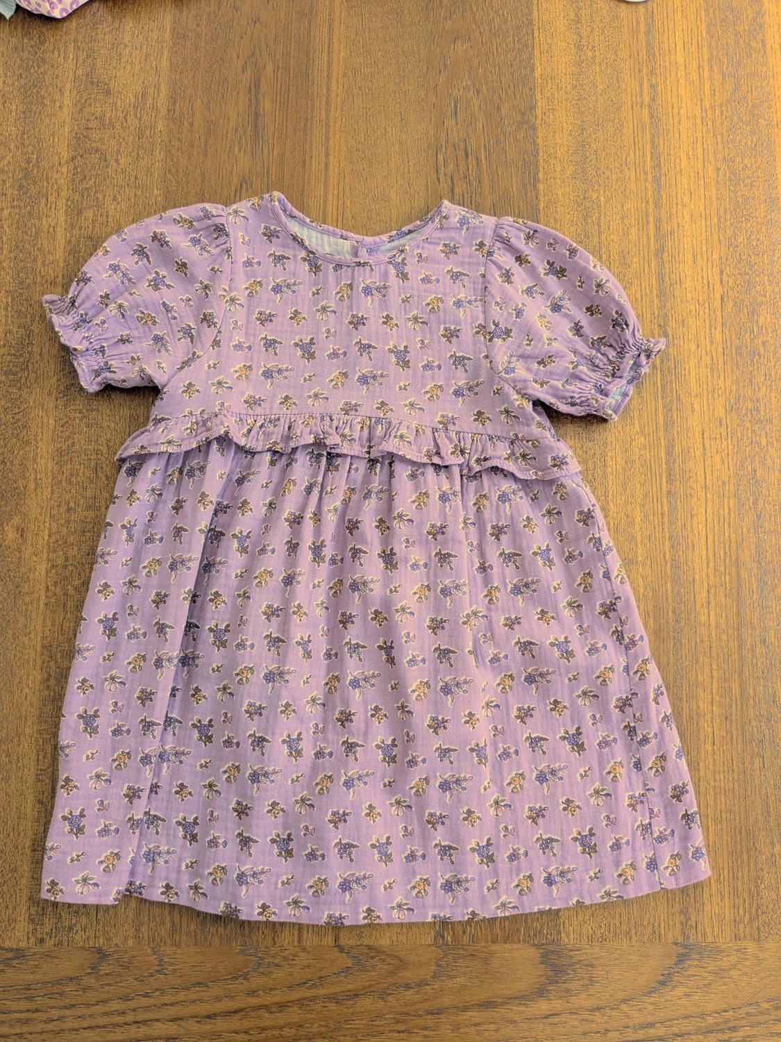 Lilac children's dress with a small floral print, puffed short sleeves, and ruffles, on a wooden table.