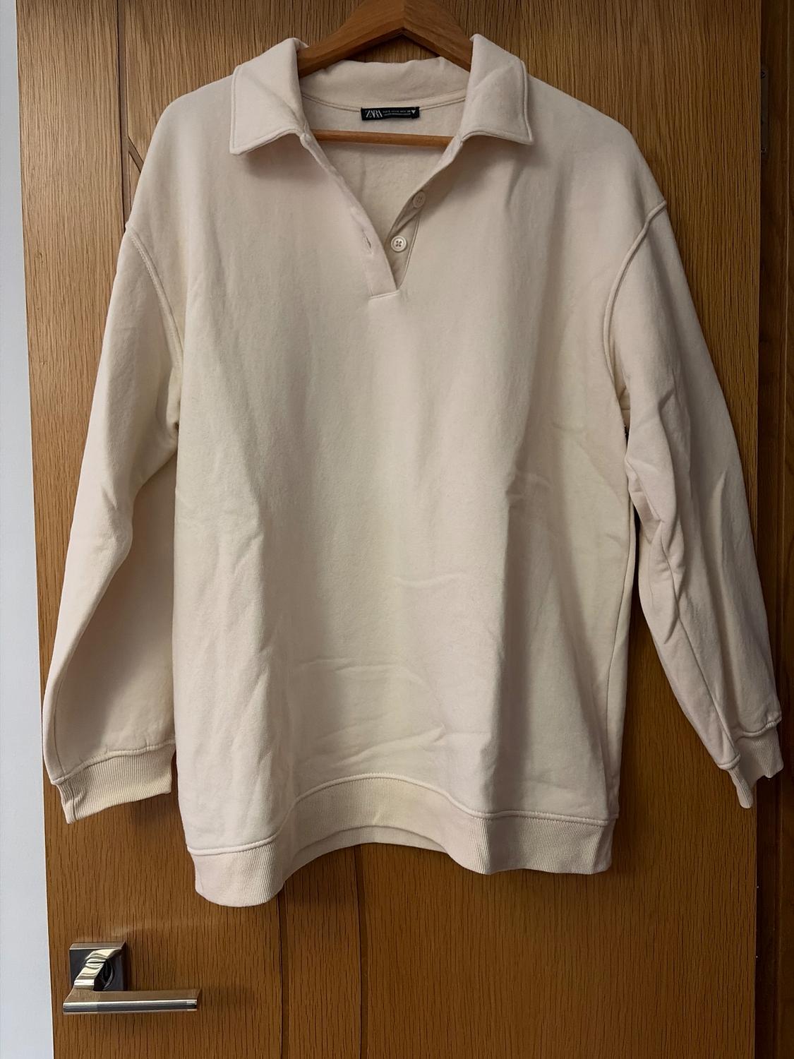 Beige long-sleeved polo-neck jumper with buttons, hanging on a wooden hanger over a light wood door.