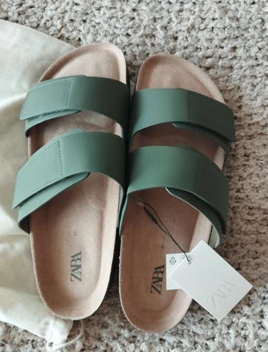 Green two-strap sandals on a beige rug, with a white ZARA label. Light fabric bag next to them.