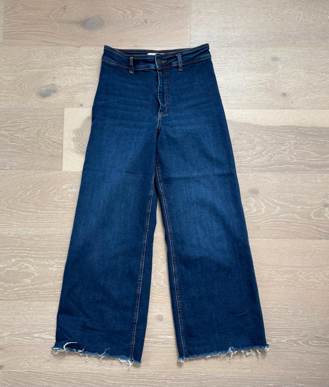 Dark blue denim pants, with a relaxed fit and a high waist, with frayed hems, placed on a light wood floor.