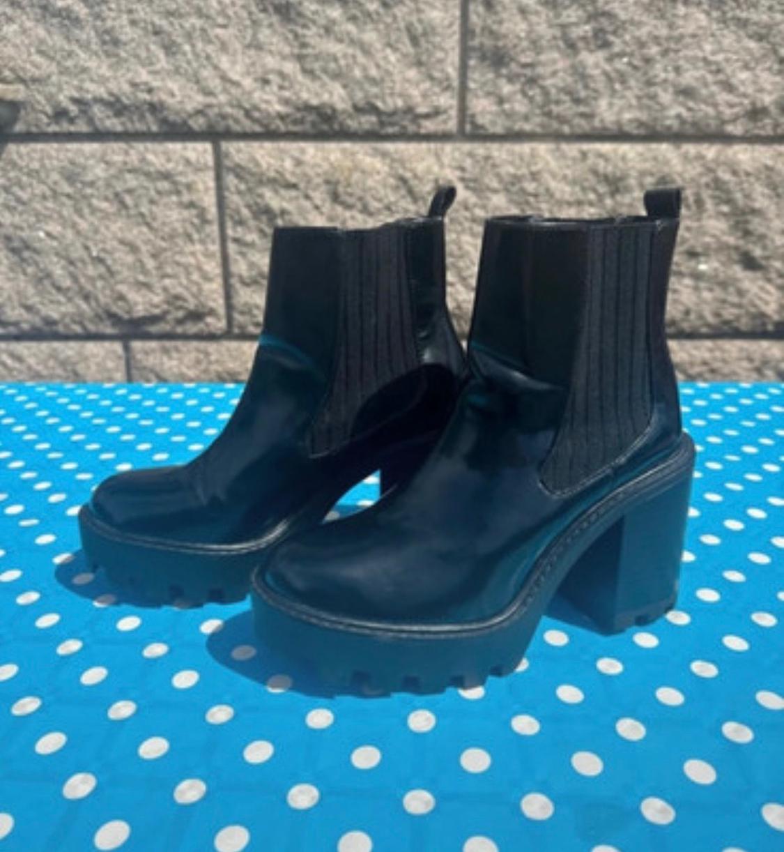 Black chunky-heeled ankle boots on a blue table with white polka dots, grey brick wall background.
