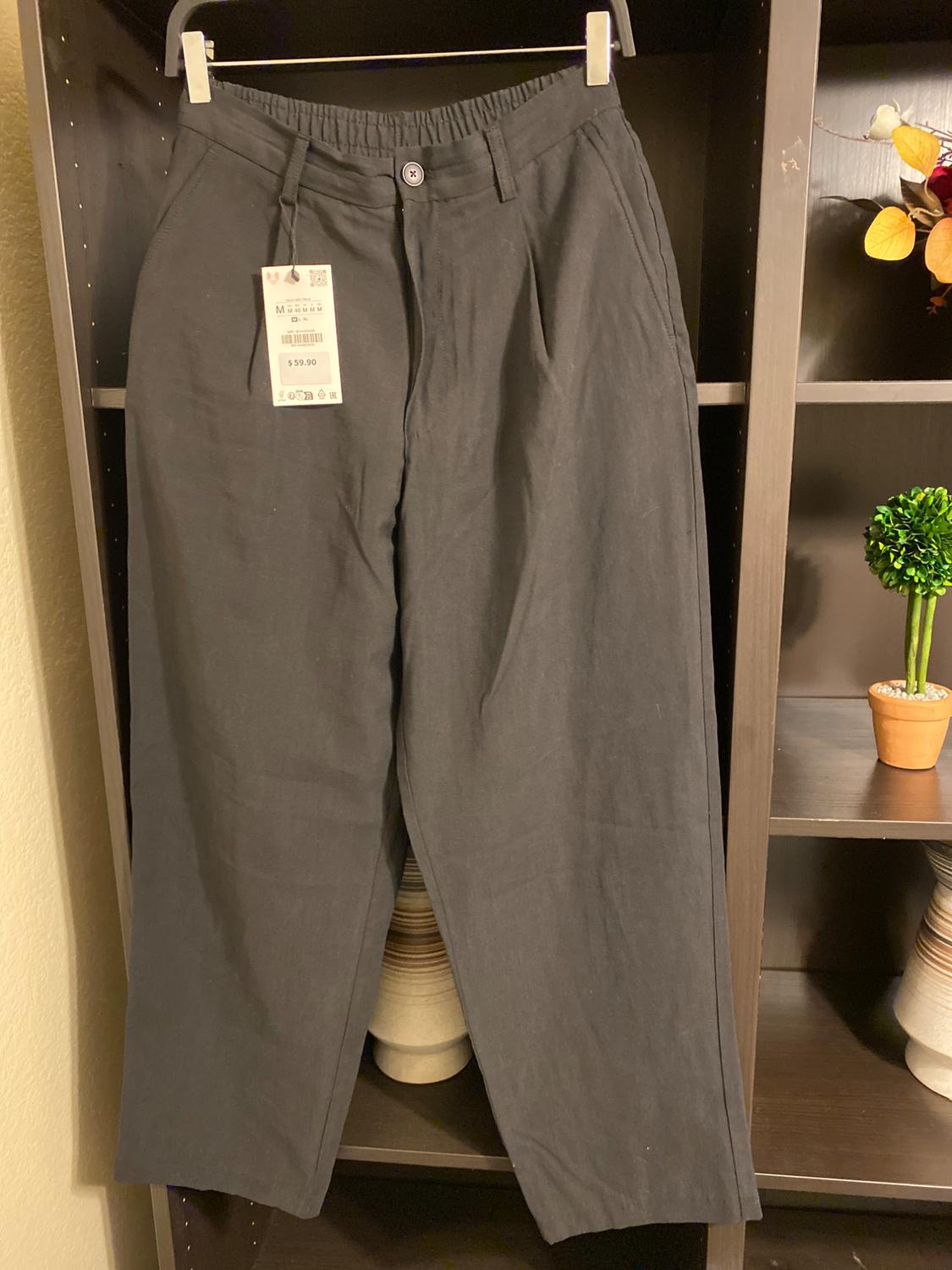 Black trousers hanging on a hanger with a price tag of $59.90, against a background of shelving with minimalist decor.