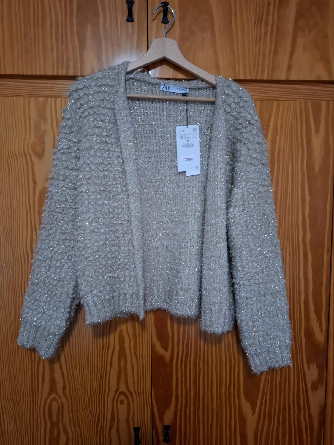 Textured beige cardigan hanging on a wooden hanger, with a visible label, against a background of wooden doors.