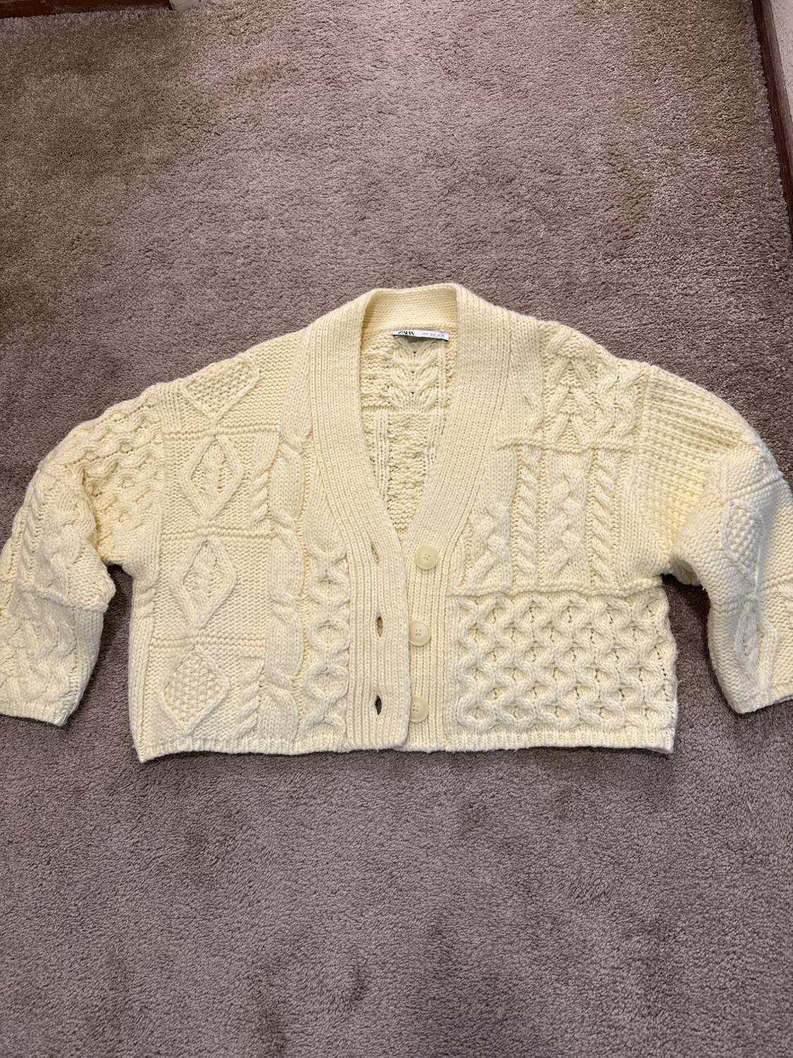 Cream-colored chunky knit cardigan with cable-knit texture and front buttons, on a brown rug.