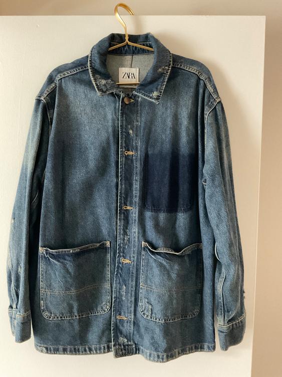 Blue denim jacket with large pockets and "ZARA" label on the collar, hanging on a golden hanger against a white background.