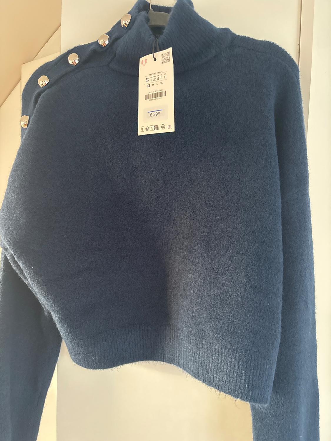 Dark blue jumper with a high neck and metallic buttons on the shoulder. Visible tag with a price of €39.95.