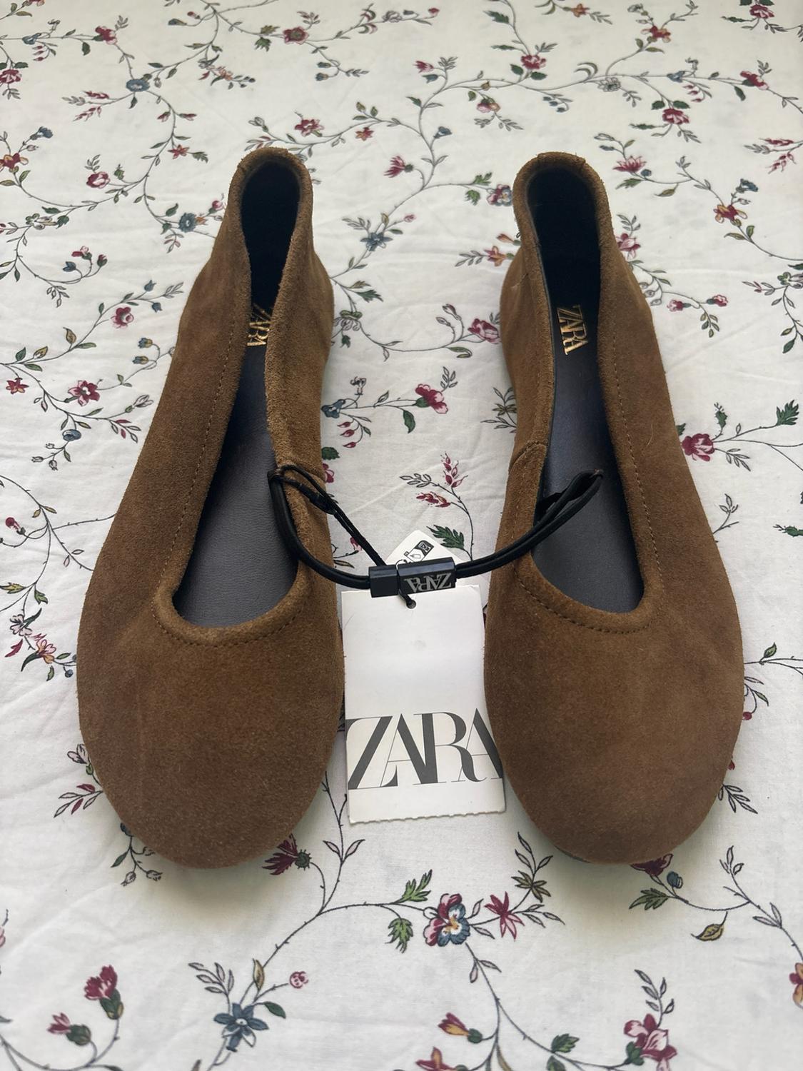 Brown suede flat shoes on a floral-print fabric background. They have a label with the text Zara.