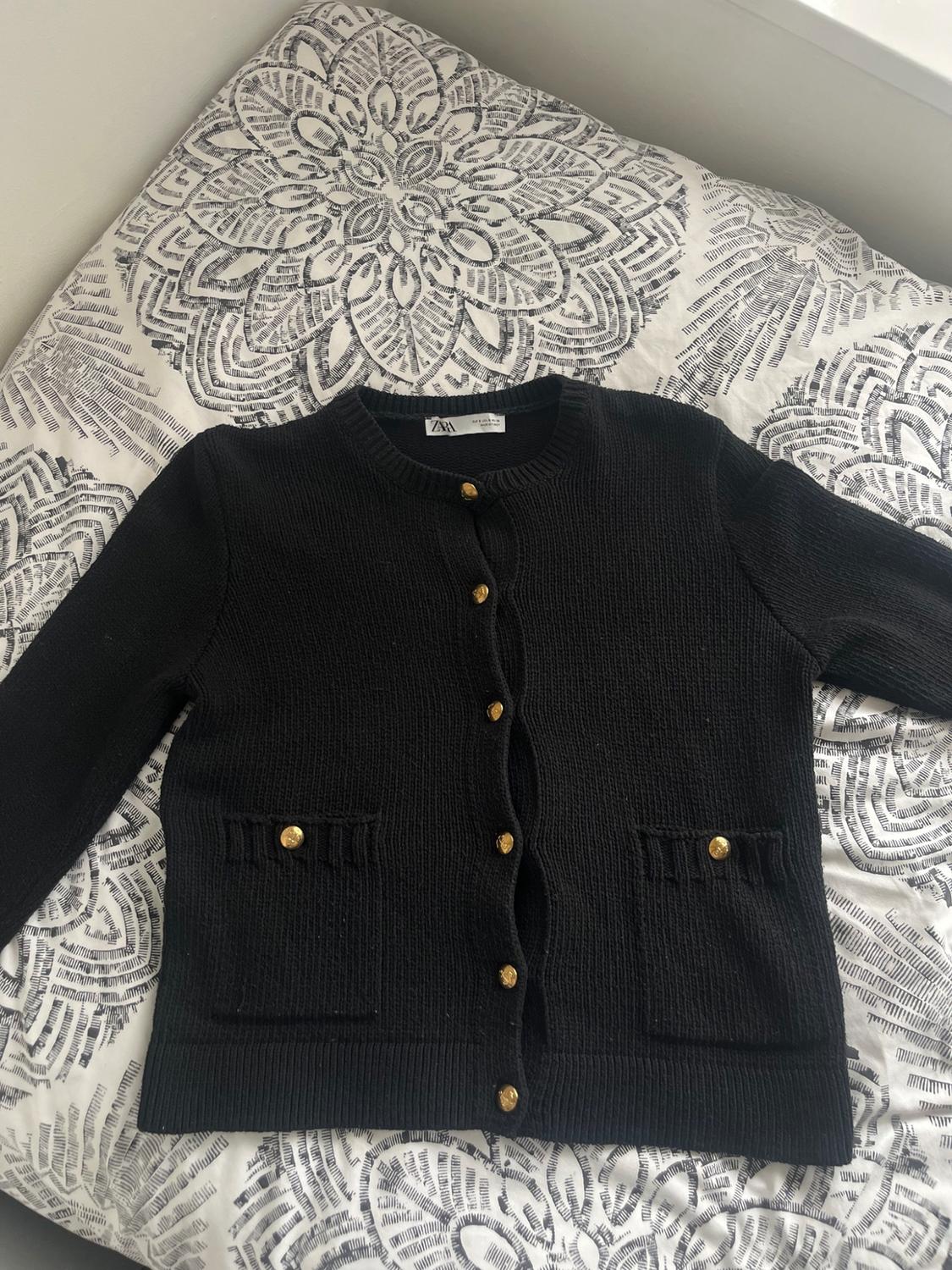 Black knitted cardigan with gold buttons and front pockets, on a white and black patterned sheet. ZARA label visible.