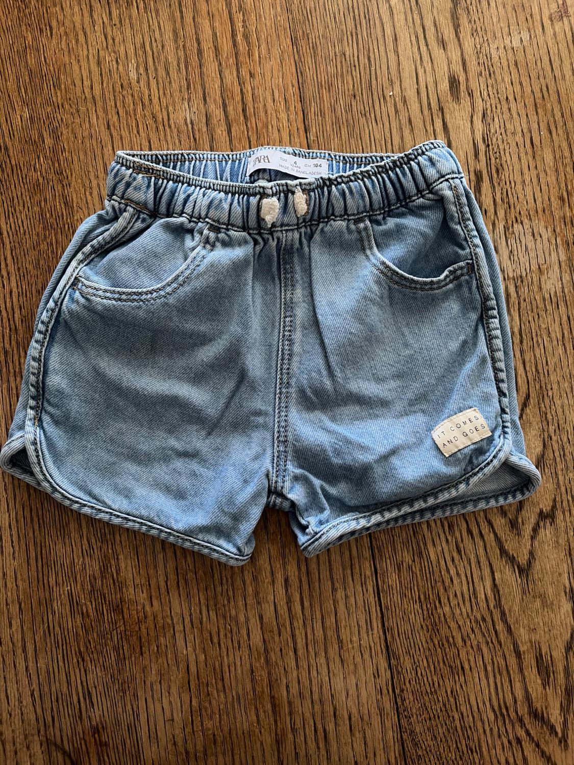 Light blue denim shorts with an elasticated waist and white drawstring, on a wooden floor. Label: "IT COMES AND GOES".