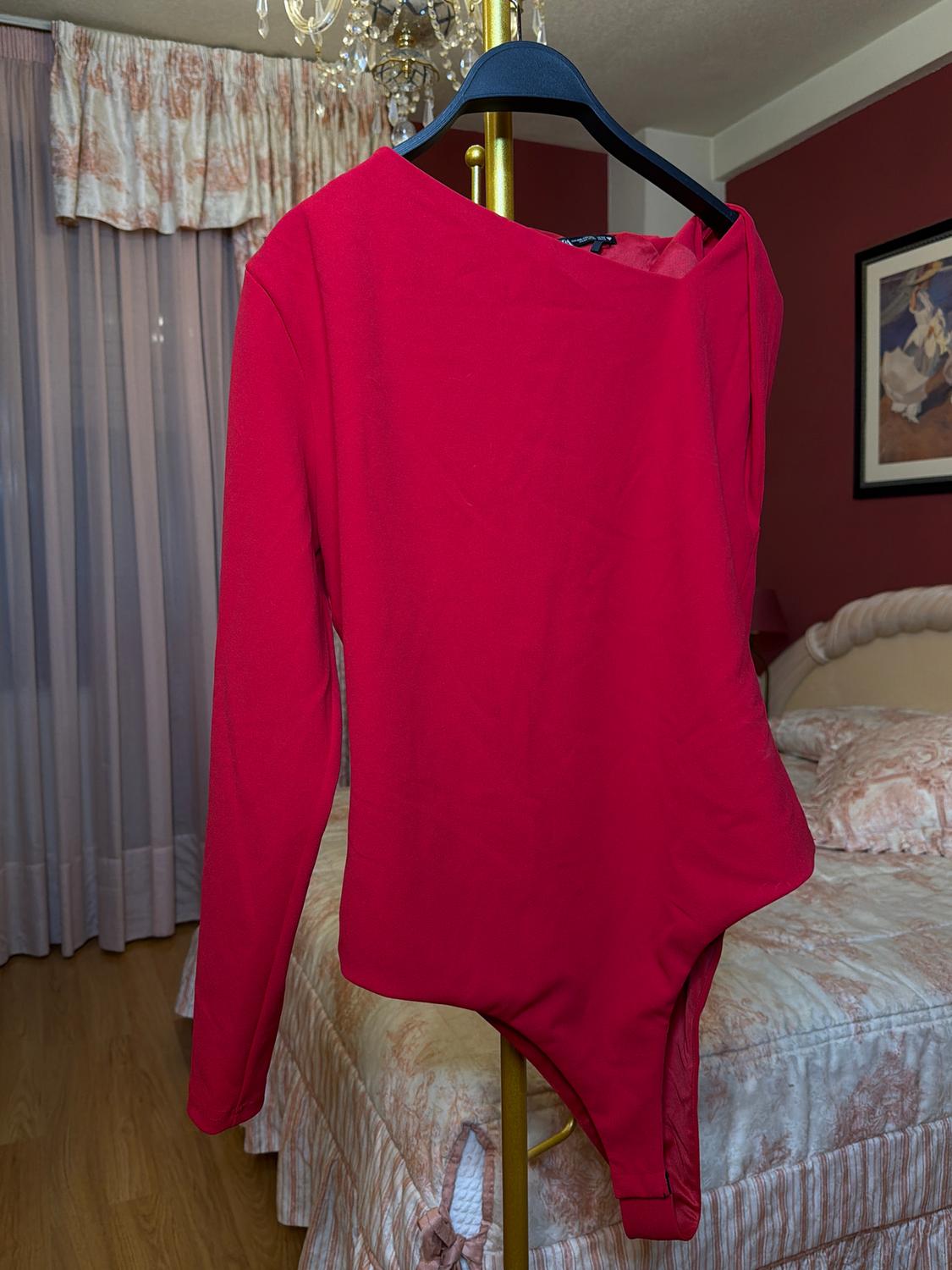 Red long-sleeved bodysuit on a black hanger, hanging in a bedroom with curtains and a bed with a light-coloured patterned bedspread.
