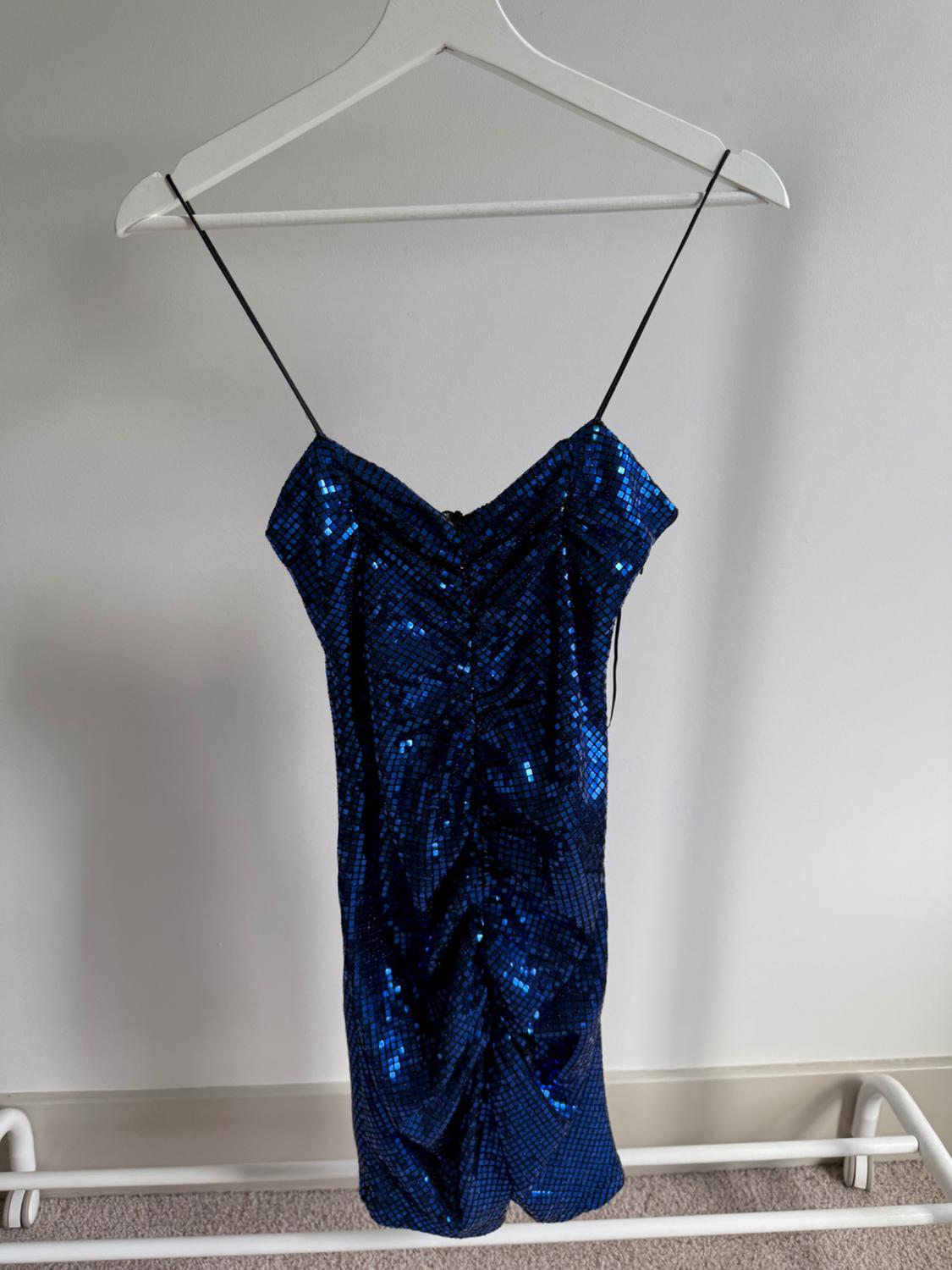 Shiny blue dress with sequins, thin black straps, hanging on a white hanger against a light grey wall background.