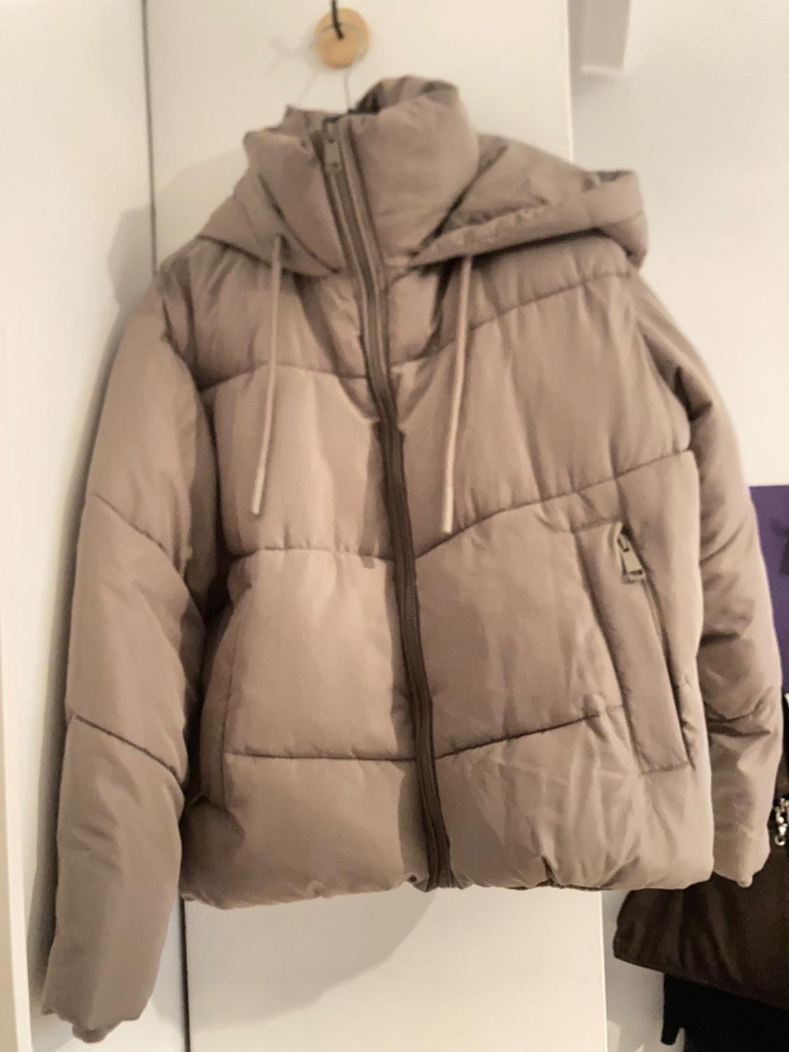 Beige padded jacket with a hood and zip fastening, hanging on a wooden hanger against a white wall.