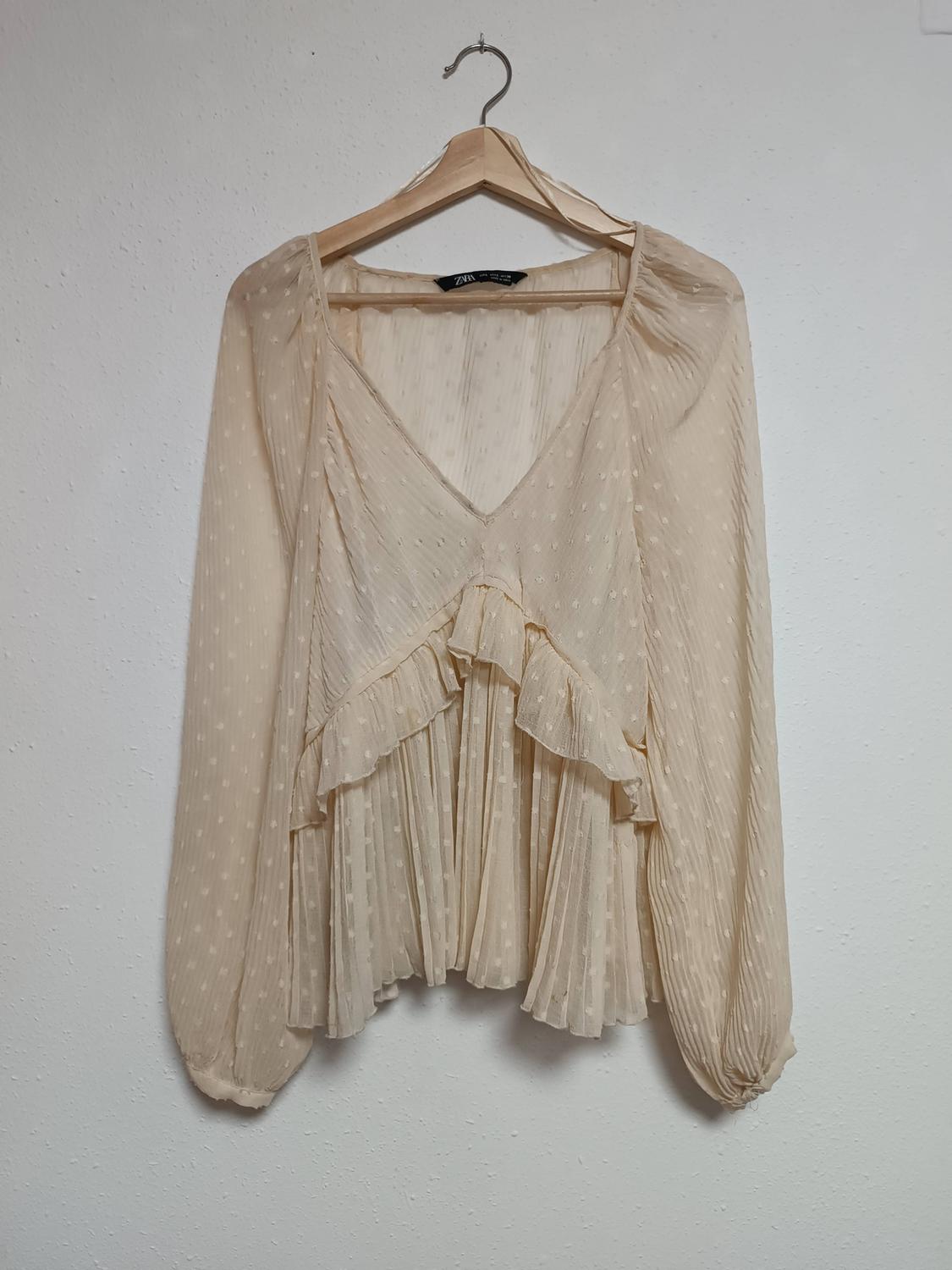 Beige chiffon blouse with long sleeves and ruffles, hanging on a wooden hanger against a white wall.