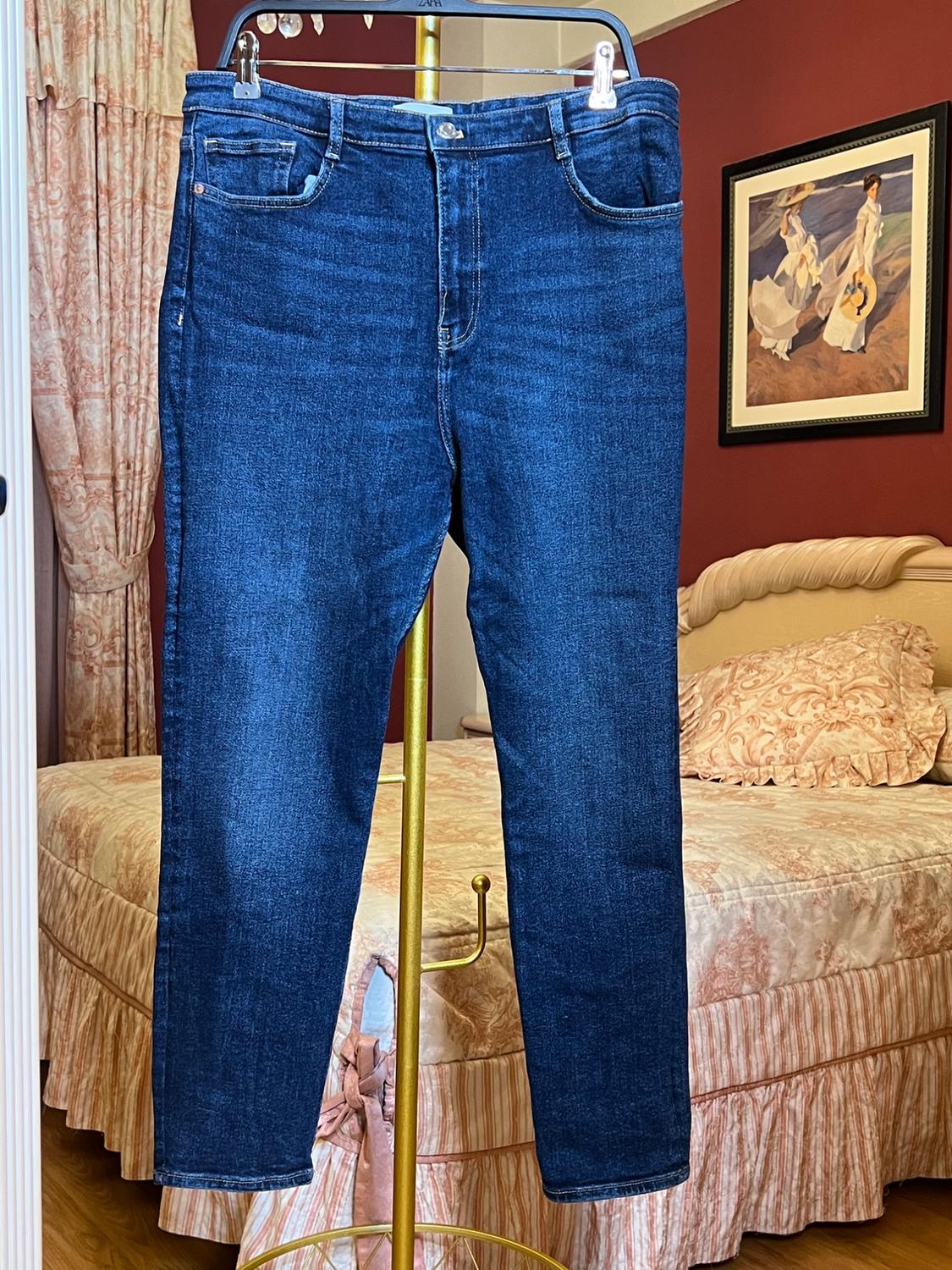Blue jeans hanging on a gold clothes rack in a bedroom with a bed and patterned curtains. Picture on the back wall.